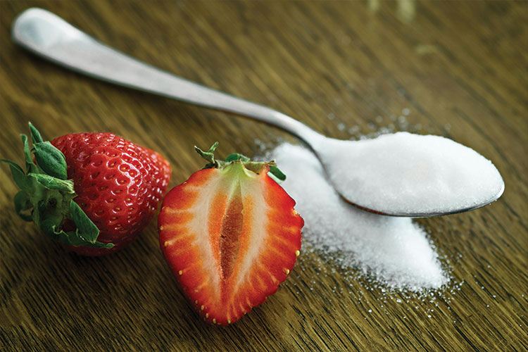 Teaspoon of white sugar next to a cut strawberry